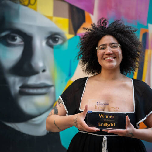 Sasha Debevec-McKenney holds a glass award in front of a colorful mural of Dylan Thomas